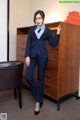 A woman in a blue suit leaning against a dresser.